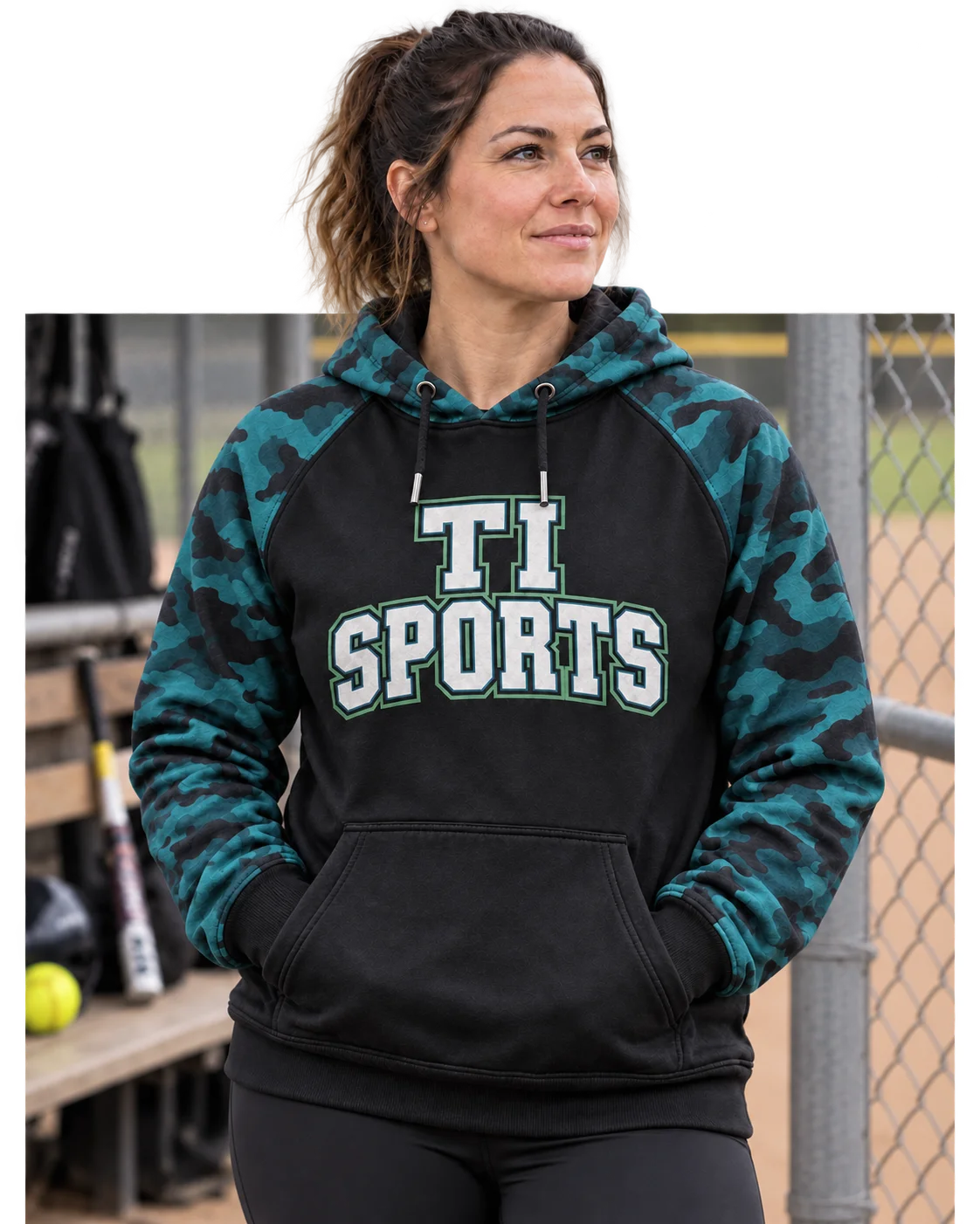Apparel concept: a woman in a charcoal and teal camouflage TiSports hoodie at the softball field, stepping beyond the photo frame